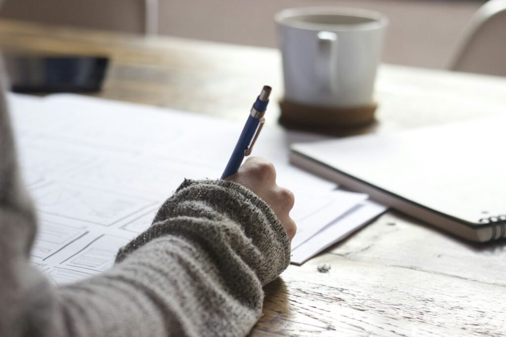 Person writing study notes by hand at a wooden desk with a coffee mug