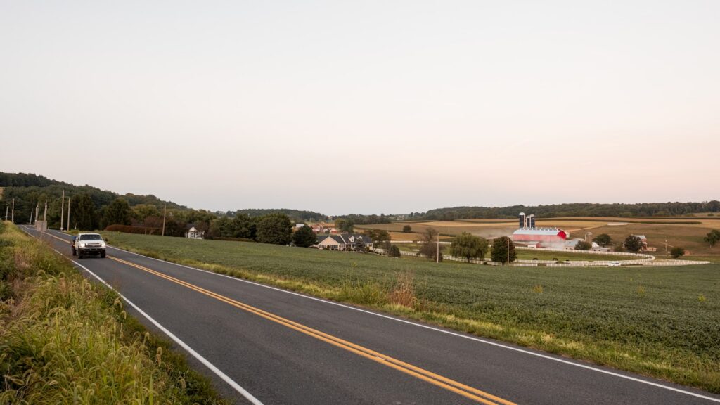 A car drives down a rural two-lane road past open fields