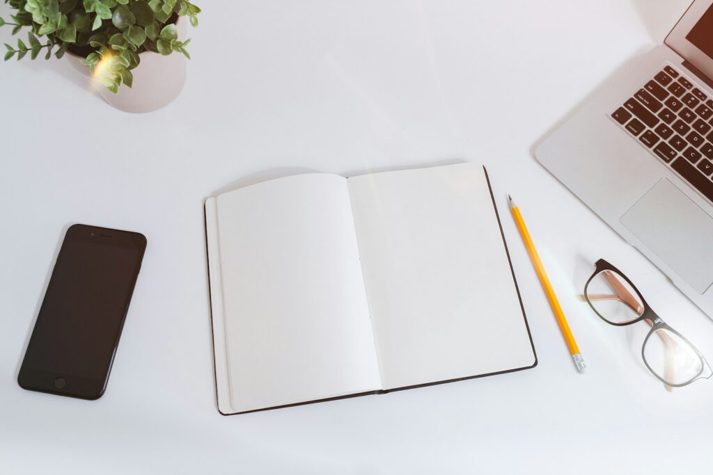 An open notebook on a white desk beside a smartphone and a laptop