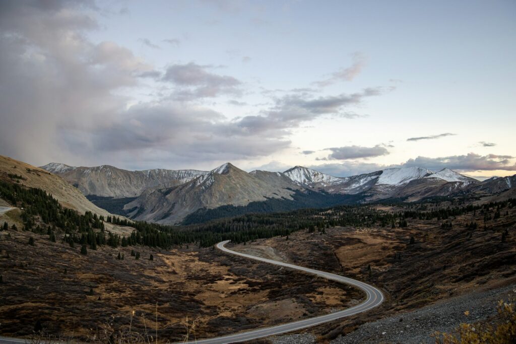 A paved two-lane road curving through the Colorado mountains under a cloudy sky
