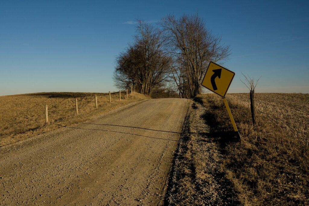 Yellow diamond shaped curve warning sign standing beside a gravel road that bends out of sight over a rise