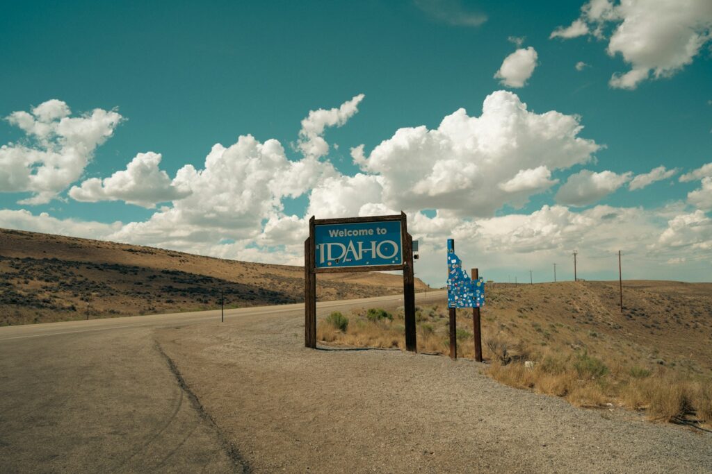 A Welcome to Idaho state line sign standing beside an empty desert highway under cumulus clouds