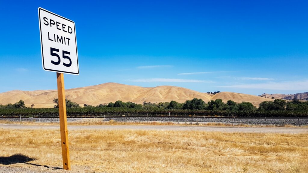 A white SPEED LIMIT 55 sign beside a two-lane road with dry golden hills and a vineyard behind it