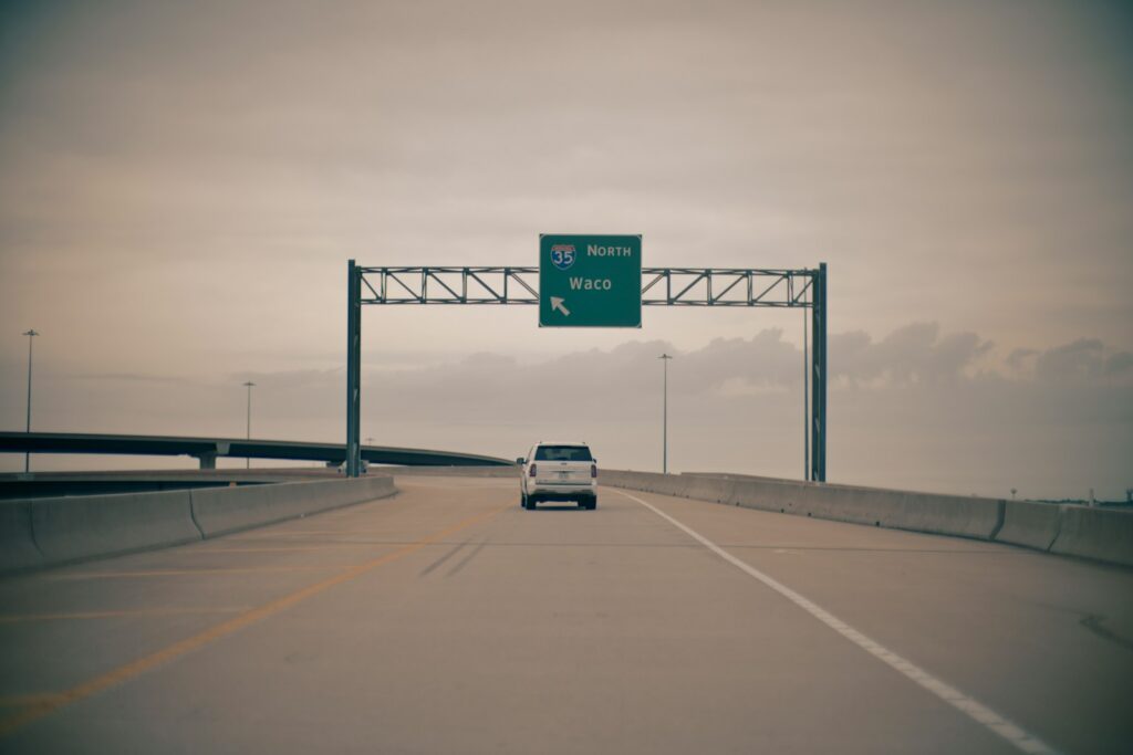 White SUV on an elevated concrete interstate ramp approaching a green overhead guide sign reading Interstate 35 North toward Waco under an overcast sky