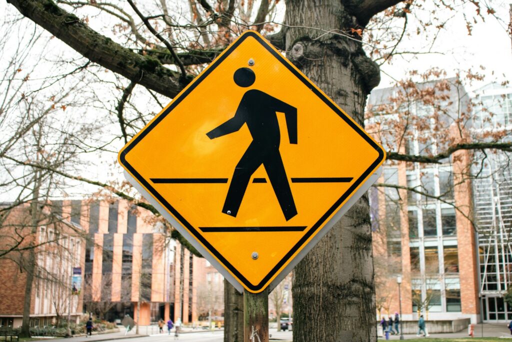 Yellow diamond pedestrian crossing sign mounted on a post beside a tree-lined city street