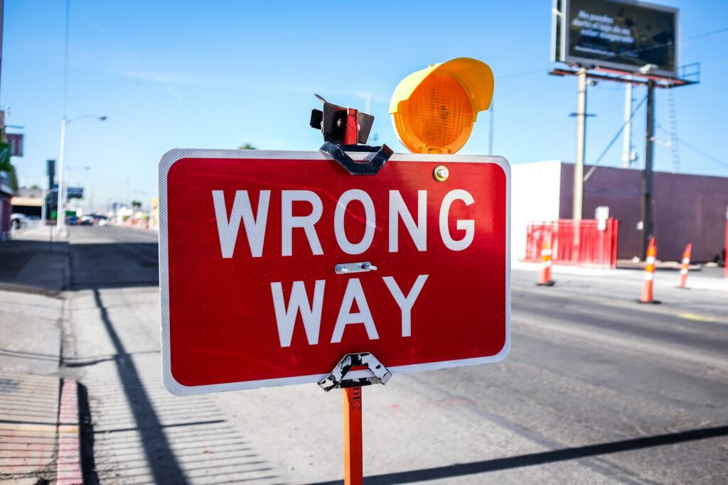A red and white rectangular WRONG WAY sign mounted at the edge of a paved road under a clear sky