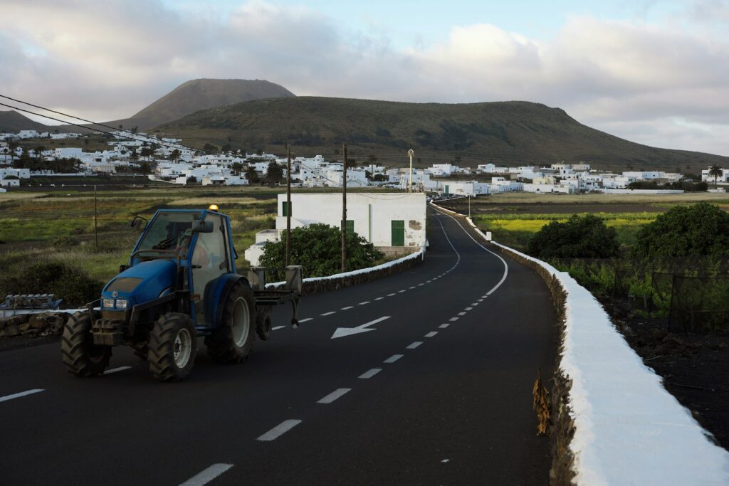 Blue tractor with an amber beacon travelling on a paved two-lane road toward a village