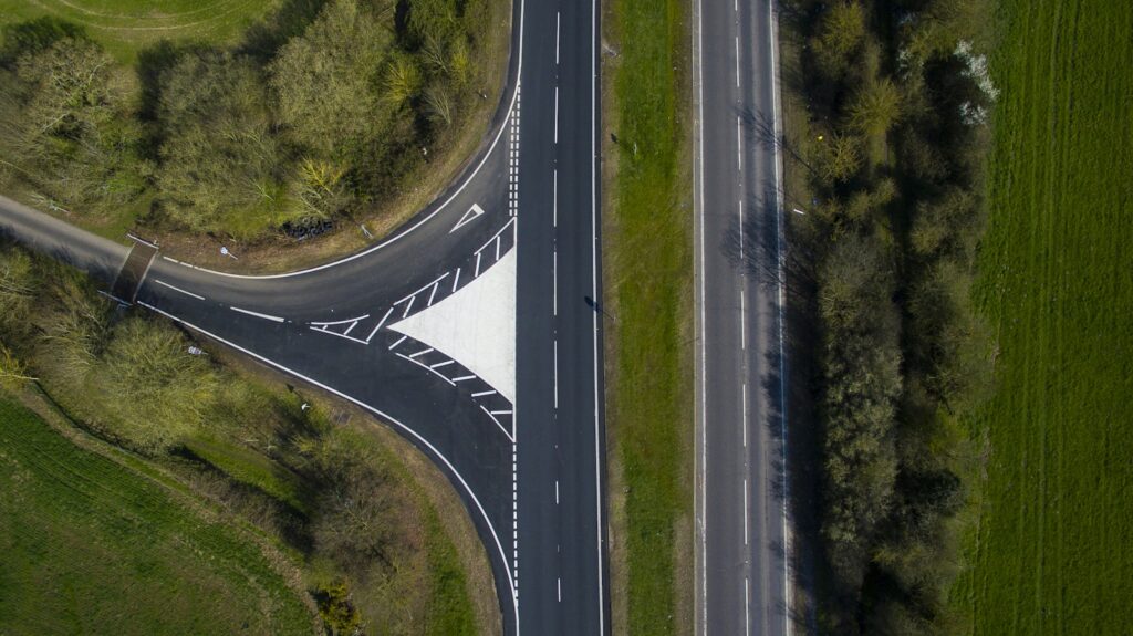 Aerial view of a side road ending at a through road in a T junction, with white give-way triangles painted on the pavement and green fields on both sides