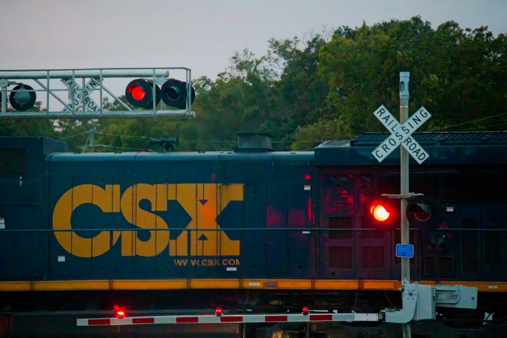 A CSX freight locomotive passing a railroad crossing at dusk with the gate lowered, red signal lights flashing, and a white crossbuck sign beside the road