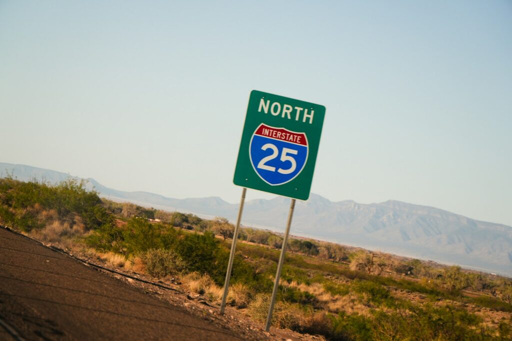Green Interstate 25 north highway sign beside a desert road with mountains behind it