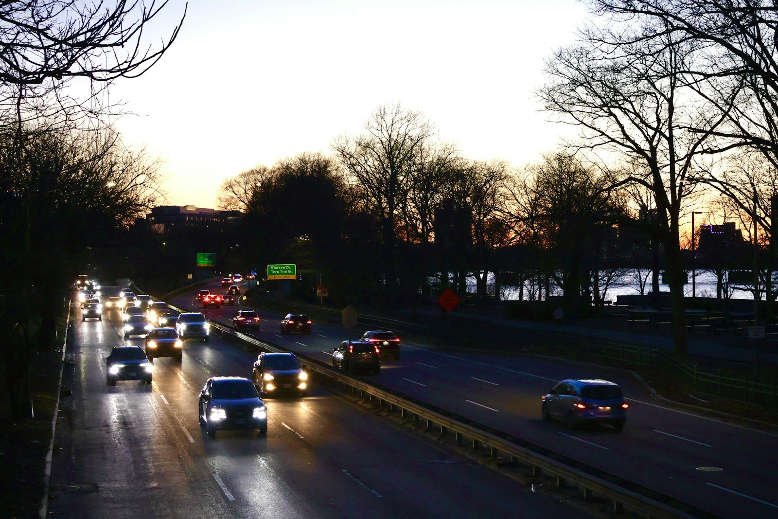 Evening traffic on Storrow Drive in Boston with headlights on at dusk, the Charles River and a green Storrow Drive thru traffic guide sign visible