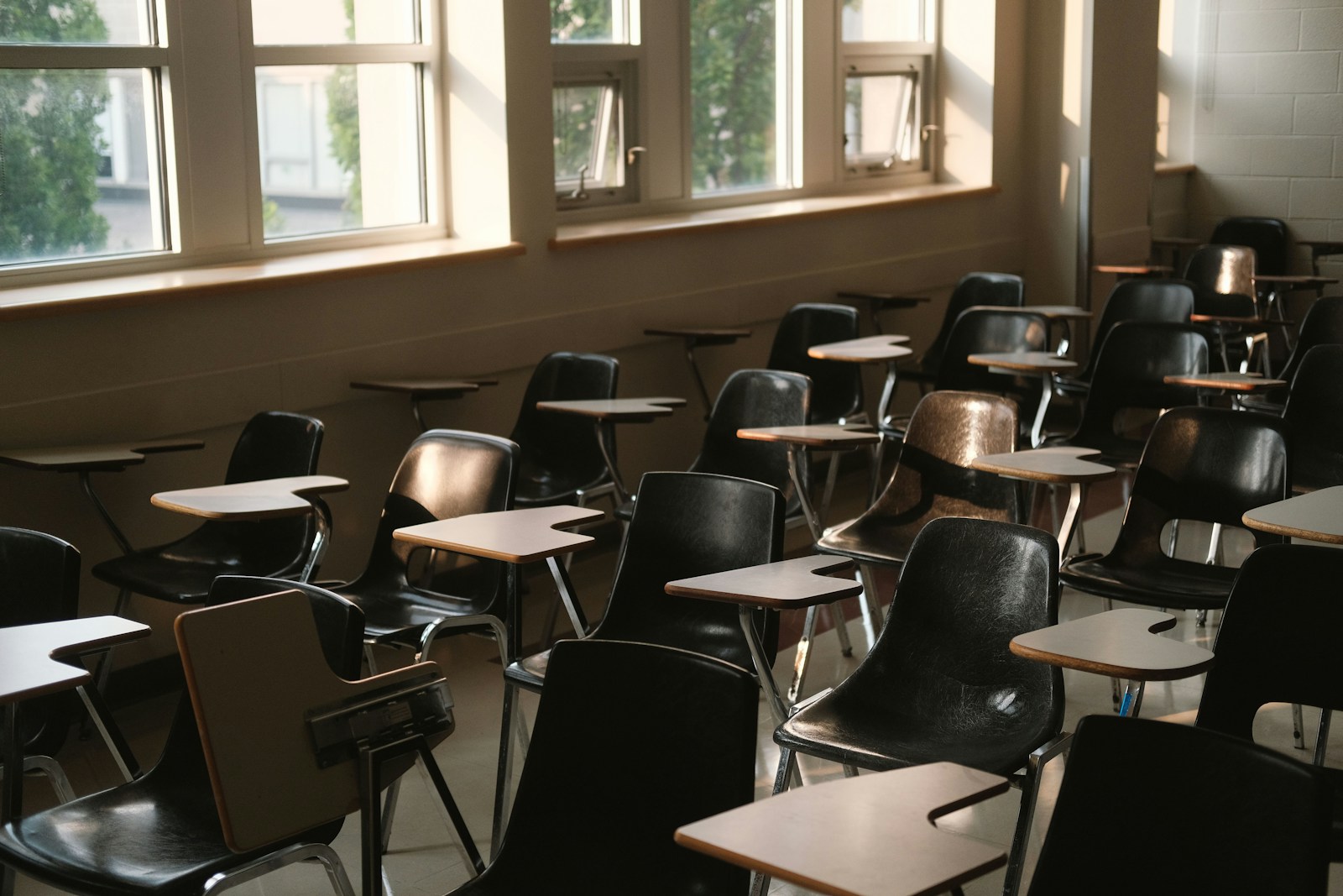 Rows of empty tablet-arm classroom chairs beside tall windows in a US school room